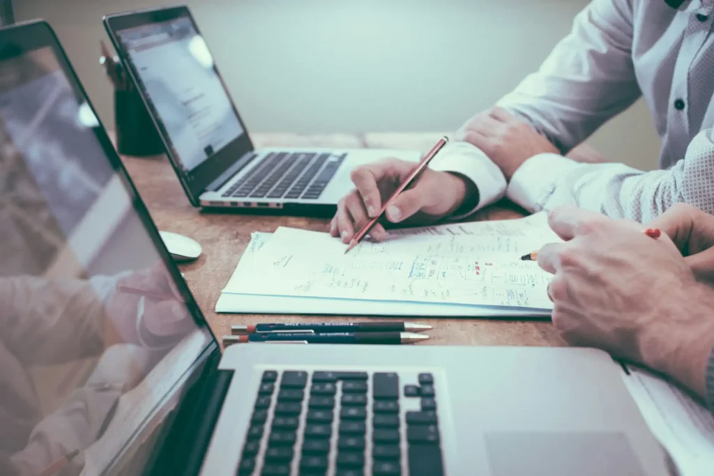 In the picture, two people working: we can see their hands holding a pen and some papers. In front of them, two laptops.